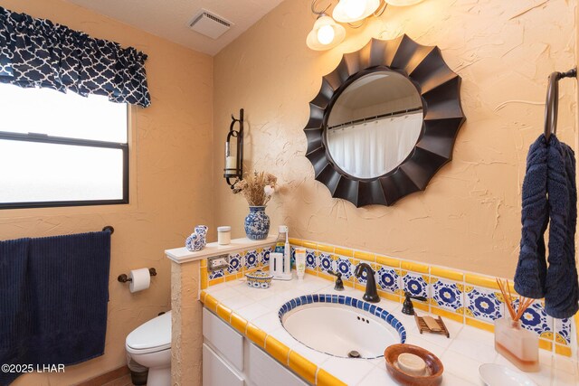 bathroom with toilet, a textured wall, vanity, and visible vents