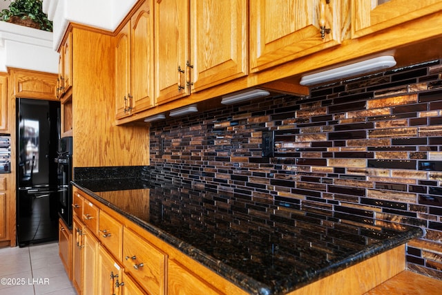 kitchen featuring tasteful backsplash, brown cabinetry, light tile patterned flooring, dark stone countertops, and black appliances