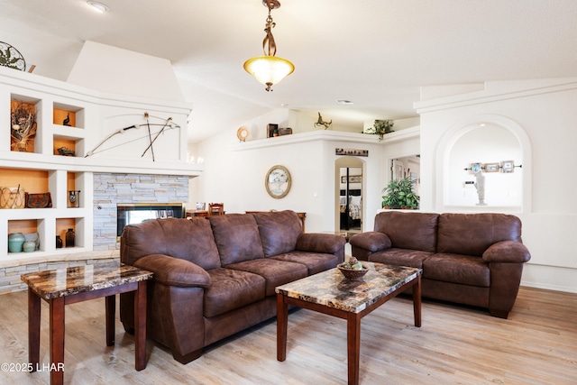 living area featuring arched walkways, a stone fireplace, built in shelves, vaulted ceiling, and light wood-type flooring