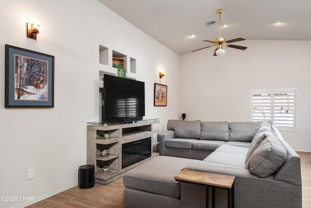 living area featuring visible vents, a ceiling fan, a glass covered fireplace, vaulted ceiling, and wood finished floors