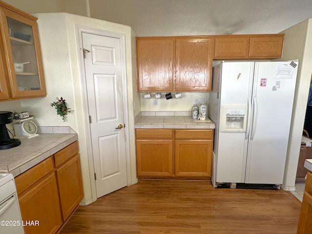 kitchen with light wood-style floors, tile counters, white refrigerator with ice dispenser, and glass insert cabinets