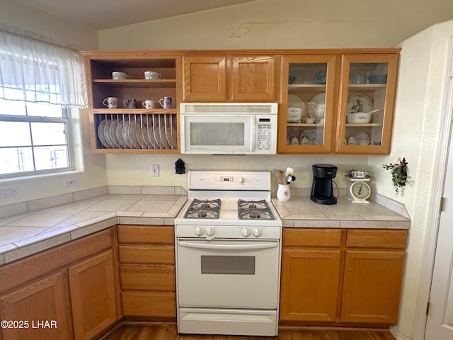 kitchen featuring white appliances, tile countertops, glass insert cabinets, brown cabinets, and vaulted ceiling