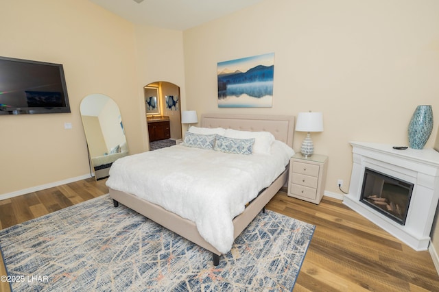 bedroom featuring arched walkways, wood finished floors, a glass covered fireplace, and baseboards