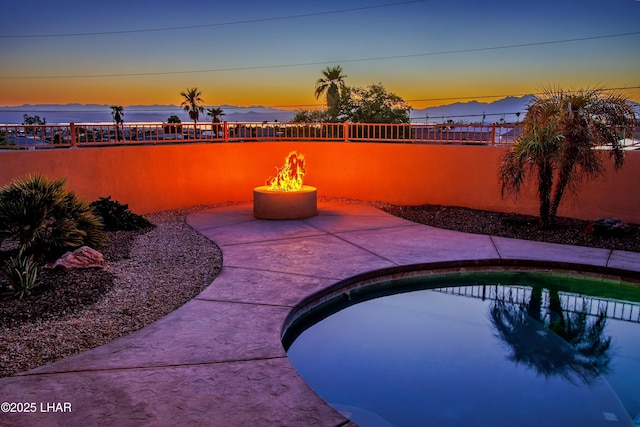 pool at dusk with a water view, an outdoor fire pit, a patio area, fence, and a swimming pool