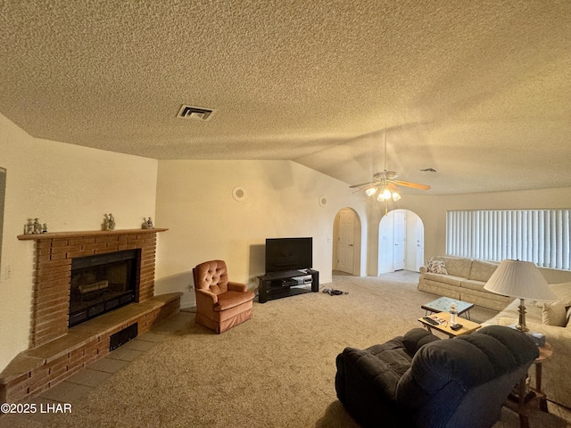 carpeted living area with arched walkways, ceiling fan, a textured ceiling, vaulted ceiling, and a brick fireplace