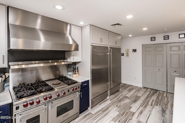 kitchen with blue cabinetry, high end appliances, white cabinetry, light hardwood / wood-style floors, and wall chimney range hood