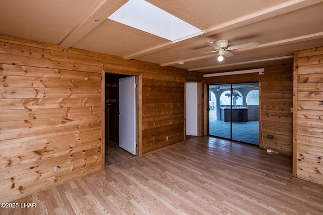 spare room with light wood finished floors, beamed ceiling, wood walls, a skylight, and a ceiling fan