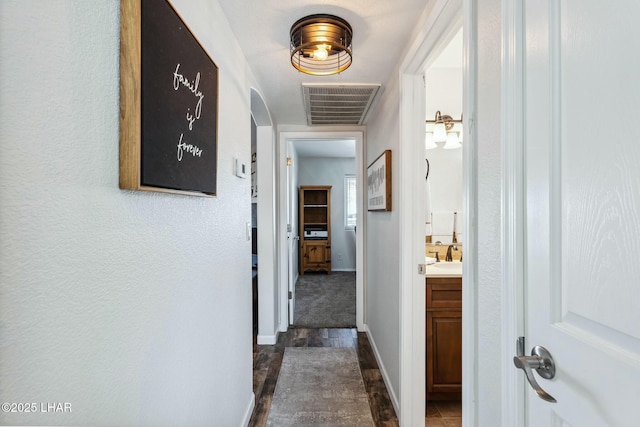 hallway featuring visible vents, a sink, arched walkways, baseboards, and dark wood-style flooring