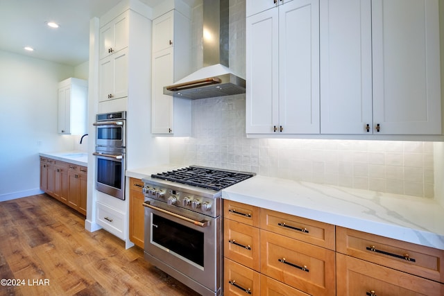 kitchen featuring white cabinetry, light stone counters, stainless steel appliances, light wood-type flooring, and wall chimney exhaust hood