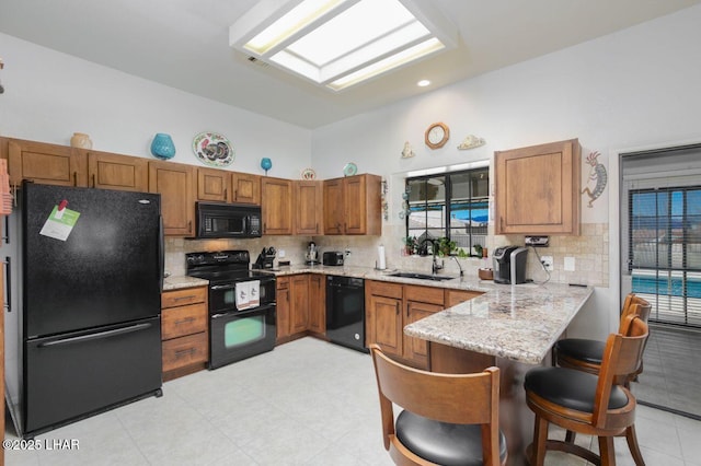 kitchen featuring brown cabinetry, a peninsula, light stone countertops, black appliances, and a sink