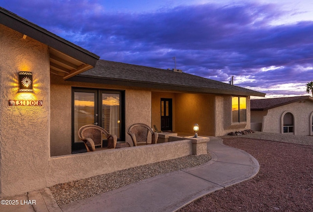 back of house at dusk with a shingled roof and stucco siding