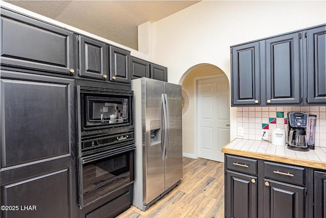 kitchen featuring arched walkways, dark cabinets, light wood-type flooring, black appliances, and tasteful backsplash