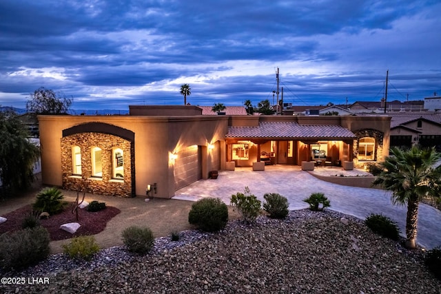 back of house at dusk with an attached garage, a tile roof, stone siding, decorative driveway, and stucco siding