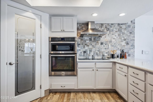 kitchen with wall chimney exhaust hood, black electric cooktop, decorative backsplash, light countertops, and double oven