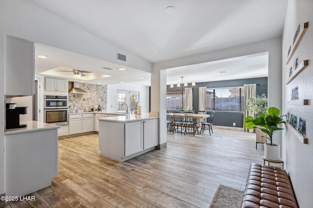 kitchen with wall chimney range hood, light wood-style floors, a healthy amount of sunlight, light countertops, and visible vents