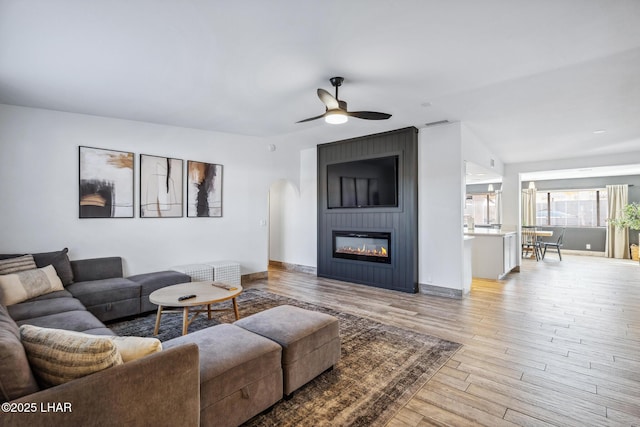 living area featuring wood finished floors, arched walkways, baseboards, a large fireplace, and ceiling fan