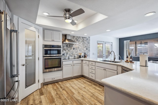 kitchen featuring light wood finished floors, a tray ceiling, a sink, wall chimney exhaust hood, and appliances with stainless steel finishes