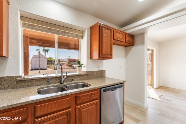 kitchen with brown cabinetry, light wood-style flooring, light stone counters, stainless steel dishwasher, and a sink