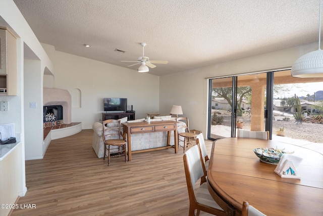 dining room with light wood finished floors, visible vents, a fireplace with raised hearth, lofted ceiling, and ceiling fan