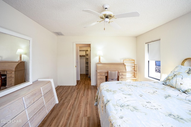 bedroom featuring a textured ceiling, wood finished floors, visible vents, a ceiling fan, and a walk in closet