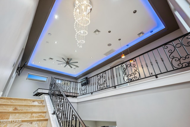 stairway with a raised ceiling, a high ceiling, ceiling fan with notable chandelier, and visible vents