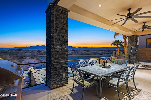 patio terrace at dusk with grilling area, outdoor dining space, a water and mountain view, and ceiling fan