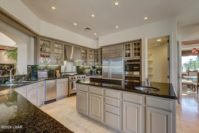 kitchen featuring a center island with sink, sink, dark stone counters, premium appliances, and wall chimney exhaust hood