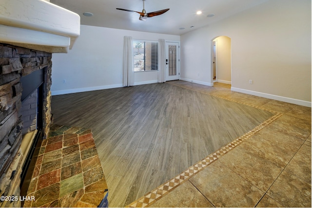 unfurnished living room with arched walkways, a fireplace with raised hearth, ceiling fan, and baseboards