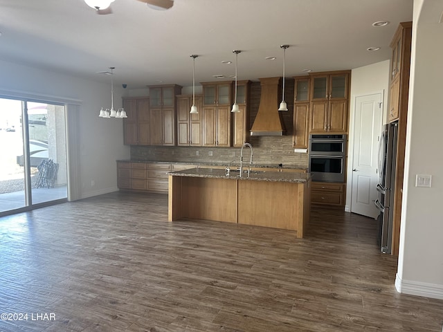 kitchen with stone counters, decorative backsplash, custom range hood, and a center island with sink