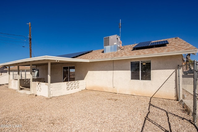 back of house featuring central air condition unit, fence, roof with shingles, roof mounted solar panels, and stucco siding