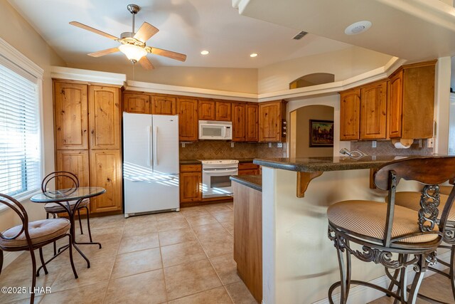 kitchen with a breakfast bar area, backsplash, light tile patterned floors, kitchen peninsula, and white appliances