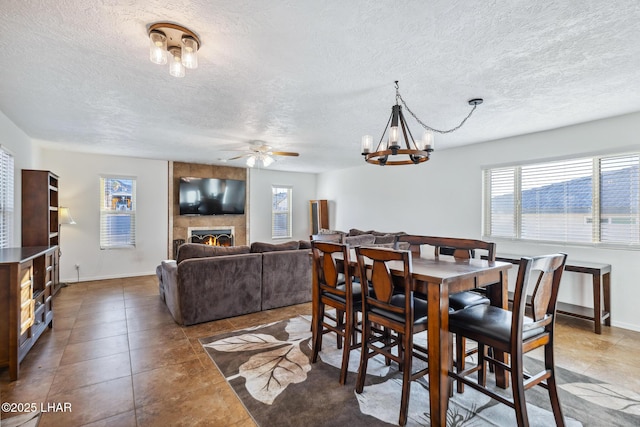 dining area with baseboards, a textured ceiling, a tiled fireplace, and ceiling fan with notable chandelier