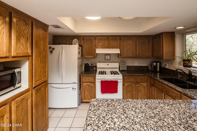 kitchen with light tile patterned flooring, sink, dark stone countertops, a tray ceiling, and white appliances