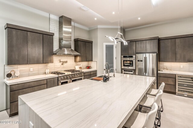 kitchen featuring wall chimney range hood, appliances with stainless steel finishes, hanging light fixtures, an island with sink, and beverage cooler