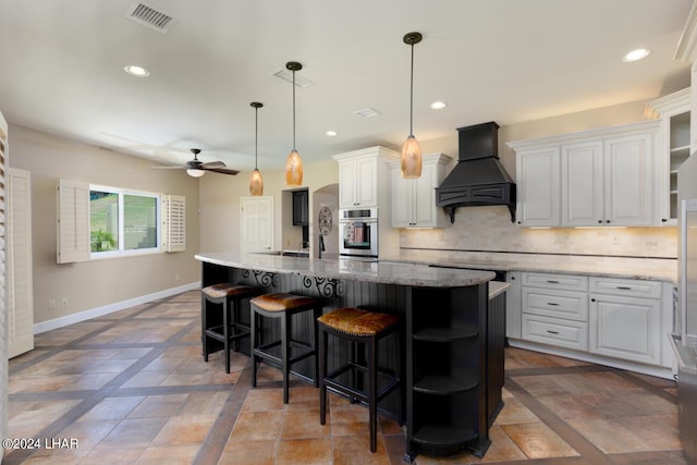 kitchen with premium range hood, white cabinetry, stainless steel oven, hanging light fixtures, and a large island with sink