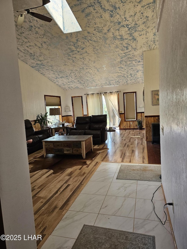 living room featuring marble finish floor, a wainscoted wall, a ceiling fan, wooden walls, and vaulted ceiling with skylight