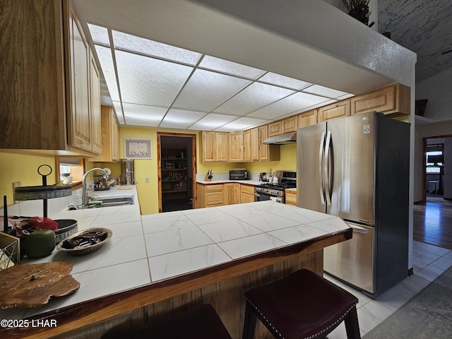 kitchen with tile countertops, a peninsula, stainless steel appliances, under cabinet range hood, and a sink