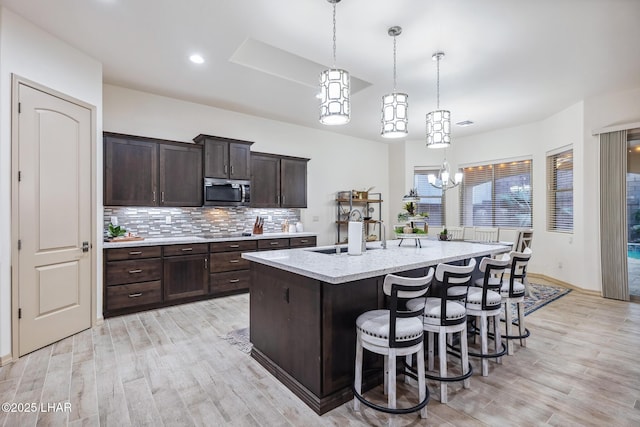 kitchen with an island with sink, a sink, stainless steel microwave, light wood-style floors, and decorative backsplash