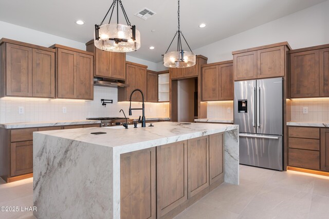 kitchen featuring an island with sink, stainless steel fridge, light stone counters, and decorative light fixtures