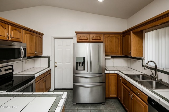 kitchen featuring a sink, tile counters, vaulted ceiling, appliances with stainless steel finishes, and brown cabinets