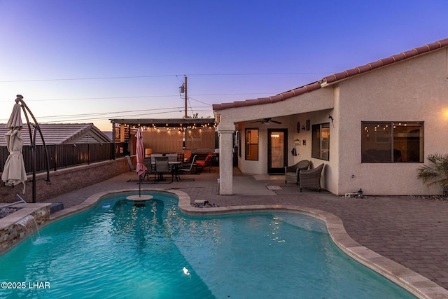 pool at dusk with pool water feature, ceiling fan, outdoor lounge area, and a patio