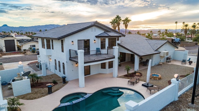 back house at dusk with a balcony, a mountain view, a pool with hot tub, and a patio area