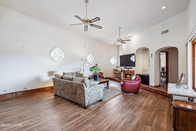 living room featuring arched walkways, ceiling fan, a high ceiling, wood finished floors, and a lit fireplace