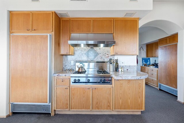 kitchen featuring light stone counters, paneled built in fridge, backsplash, wall chimney range hood, and stainless steel gas cooktop