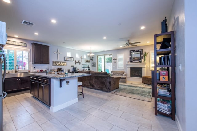 kitchen with a kitchen breakfast bar, dark brown cabinets, dark stone countertops, ceiling fan with notable chandelier, and a center island