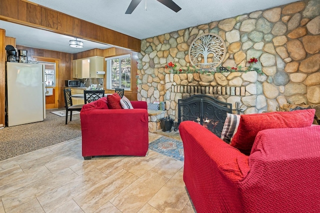 carpeted living room featuring ceiling fan, wood walls, a stone fireplace, and a textured ceiling