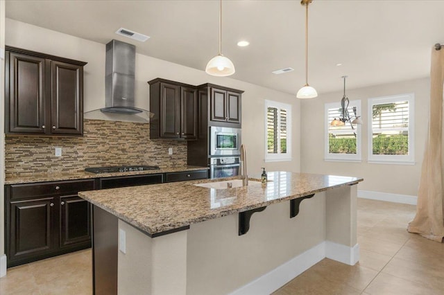 kitchen featuring light tile patterned flooring, wall chimney range hood, an island with sink, hanging light fixtures, and appliances with stainless steel finishes