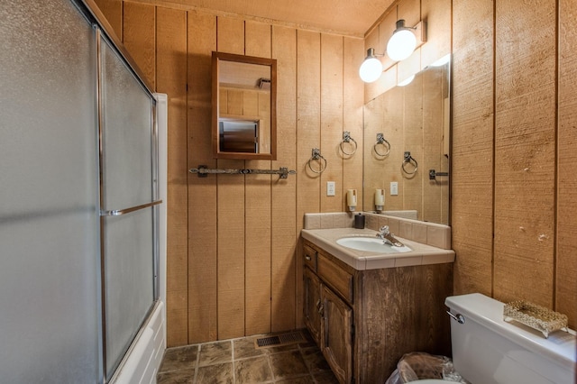 full bathroom featuring vanity, bath / shower combo with glass door, toilet, and wood walls