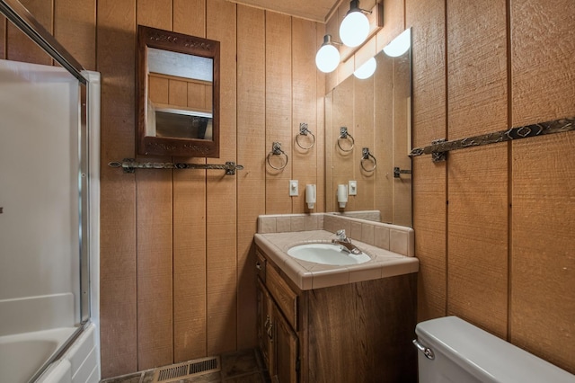 full bathroom featuring bath / shower combo with glass door, vanity, toilet, and wood walls