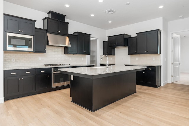 kitchen with stainless steel appliances, a center island with sink, ventilation hood, sink, and light hardwood / wood-style floors